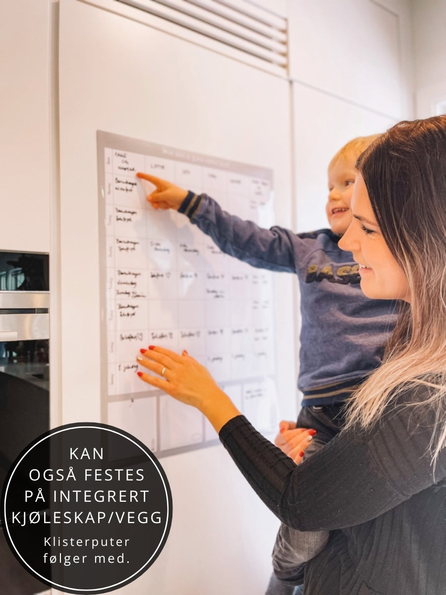 Mor og barn ser på ukens planer. Ukeplanleggeren henger på integrert kjøleskap.